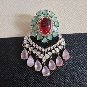 May include: An ornate earring featuring a large oval red gemstone surrounded by light blue and clear stones. The earring has a crescent shape with clear stones and dangling pink teardrop-shaped gems. The setting is gold-toned.