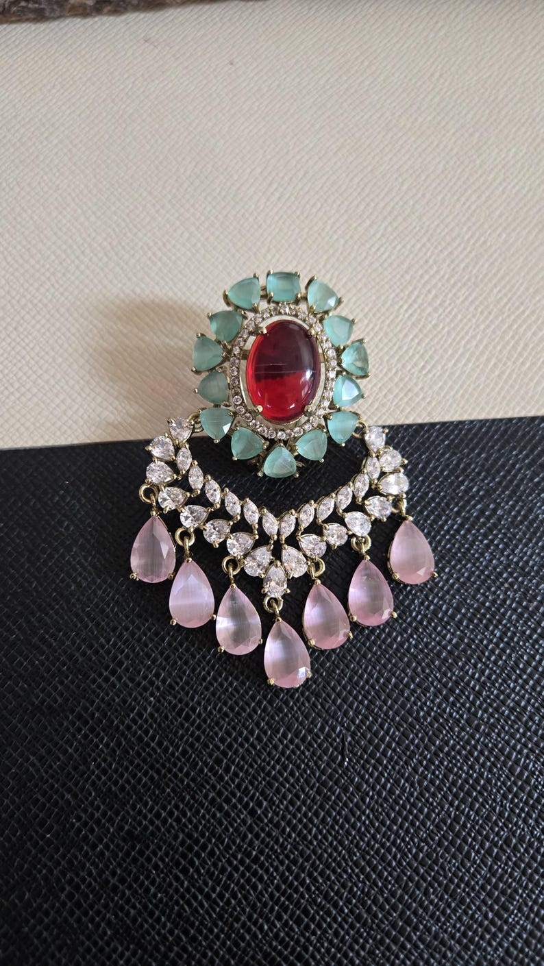 May include: An ornate earring featuring a large oval red gemstone surrounded by light blue and clear stones. The earring has a crescent shape with clear stones and dangling pink teardrop-shaped gems. The setting is gold-toned.