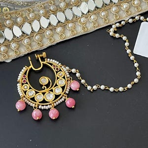 May include: A gold-toned nose ring with a circular design, adorned with clear and red stones, and pink bead dangles. The jewelry piece is attached to a delicate chain of white pearls and gold links.
