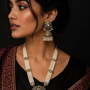 May include: A gold-toned necklace and matching jhumka earring set featuring white pearl strands and circular pendants with green enamel detailing, central clear stones, and small dangling white pearl beads throughout the jewelry pieces.