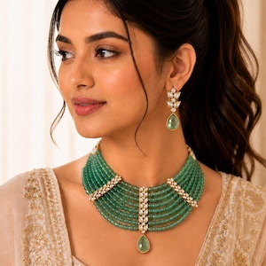 Peut inclure: Un collier ras-de-cou composé de sept rangs de petites perles vertes translucides, agrémenté de trois barrettes dorées serties de pierres blanches et d'un pendentif central en forme de goutte verte, accompagné de boucles d'oreilles assorties.