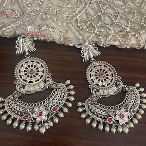 May include: Pair of silver-toned earrings with intricate filigree and red gemstone accents. The earrings feature a circular top design and a crescent-shaped bottom adorned with small pearls. The background includes a white fabric with silver embroidery.