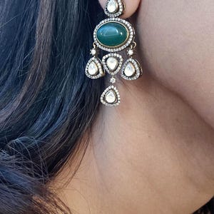 Puede incluir: Un par de pendientes colgantes con una piedra ovalada verde rodeada de piedras transparentes. Los pendientes tienen un dise&ntilde;o de candelabro con m&uacute;ltiples piedras en forma de l&aacute;grima.