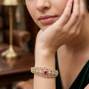 May include: A gold-toned bracelet with a central ruby and surrounding clear gemstones. The bracelet features an openwork design with a repeating pattern. The jewelry is worn on a wrist.