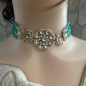 Op de afbeelding: Een gouden chokerketting met een groot, versierd bloemmotief in het midden. De ketting is versierd met witte parels en heldere stenen. De ketting ligt op een witte achtergrond.