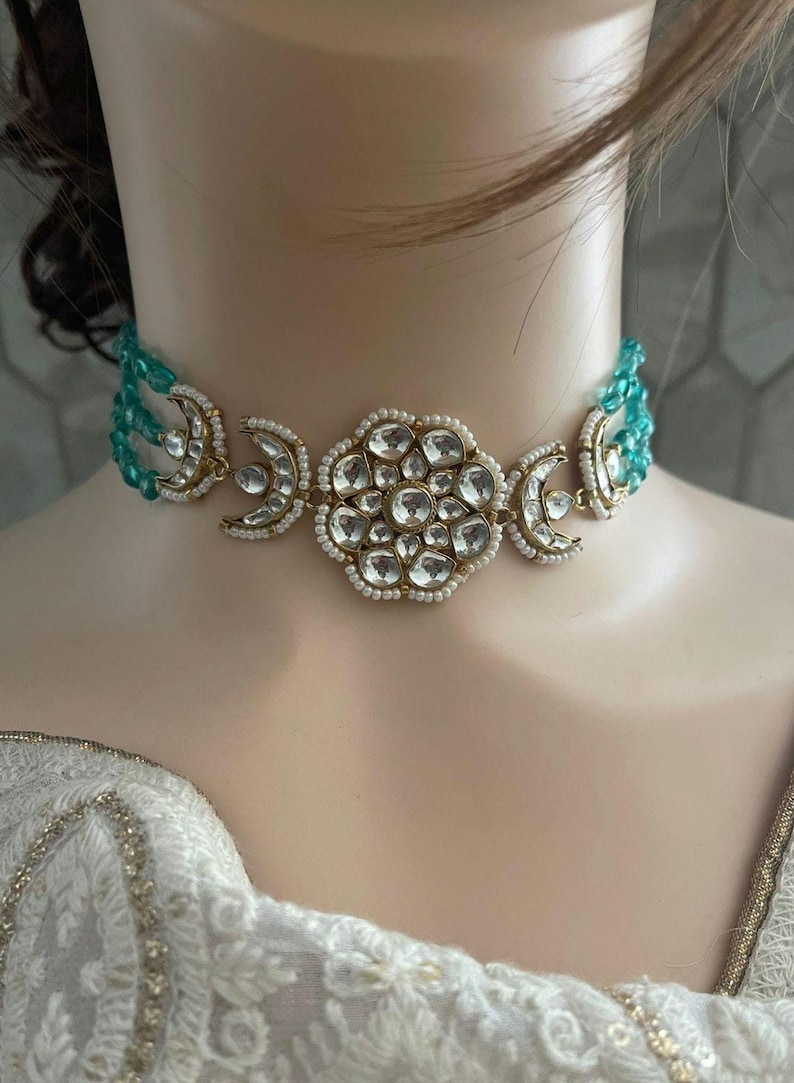 Op de afbeelding: Een gouden chokerketting met een groot, versierd bloemmotief in het midden. De ketting is versierd met witte parels en heldere stenen. De ketting ligt op een witte achtergrond.