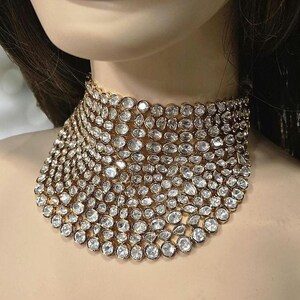 May include: A gold-coloured choker necklace, densely set with numerous clear, round-cut gemstones. The necklace is wide, covering a significant portion of the neck. The gemstones are closely arranged, creating a sparkling, luxurious appearance.
