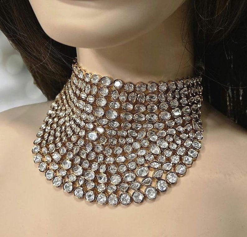 May include: A gold-coloured choker necklace, densely set with numerous clear, round-cut gemstones. The necklace is wide, covering a significant portion of the neck. The gemstones are closely arranged, creating a sparkling, luxurious appearance.