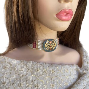 May include: A white pearl and red stone choker necklace with a gold and blue pendant featuring a floral design. The necklace is worn by a mannequin with brown hair.