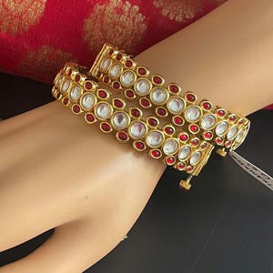 May include: Gold-toned bangles with clear and red gemstones. The bangles are adorned with alternating clear and red circular stones, set in a gold metal frame. The design is symmetrical and the bangles are displayed on a mannequin's arm.