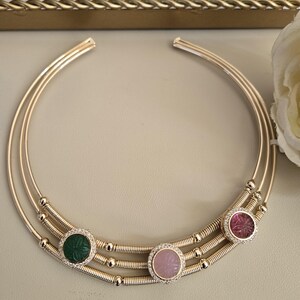 May include: Gold-toned necklace with three circular bands. The necklace features three gemstone pendants: green, pink, and red, each surrounded by a halo of small, clear stones. The necklace has a delicate, elegant design.