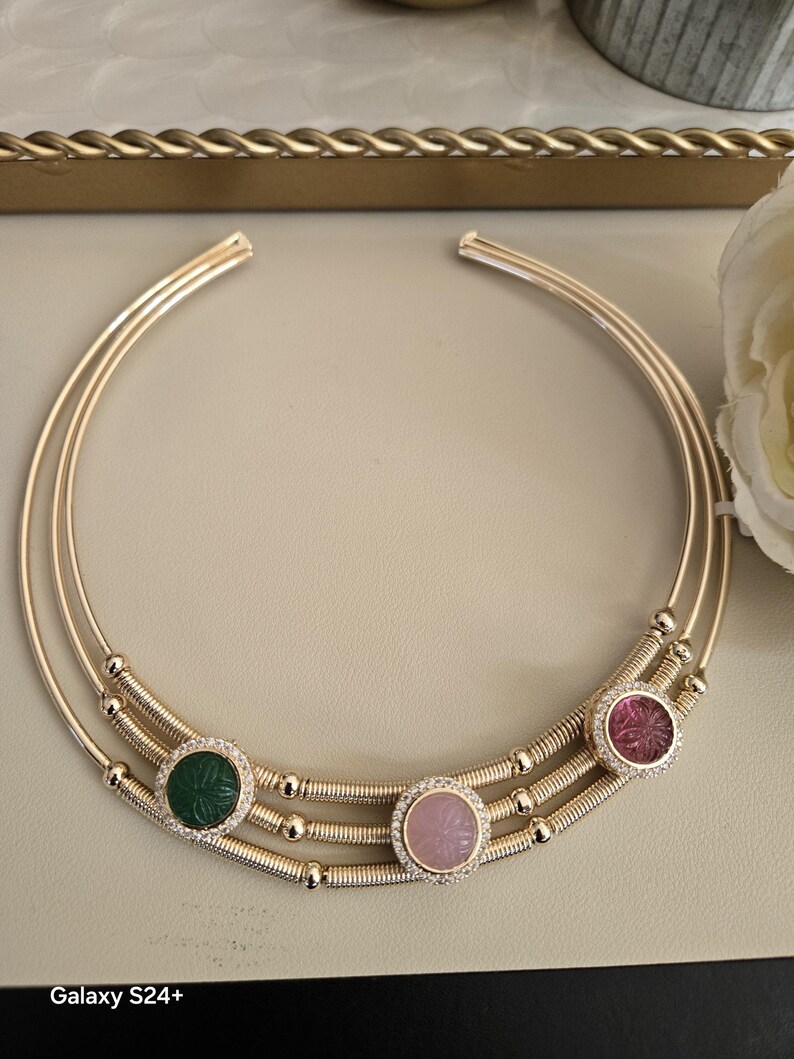 May include: Gold-toned necklace with three circular bands. The necklace features three gemstone pendants: green, pink, and red, each surrounded by a halo of small, clear stones. The necklace has a delicate, elegant design.