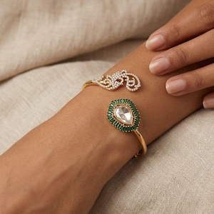 May include: Gold-toned bracelet featuring two distinct designs. One end showcases a delicate, openwork design adorned with small, clear gemstones. The other end features a teardrop-shaped, clear gemstone surrounded by emerald-green stones. The bracelet is worn on a wrist.