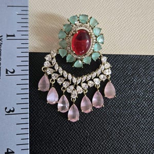 May include: An ornate earring featuring a large oval red gemstone surrounded by small, light green stones. Below, a crescent of clear, sparkling stones and dangling pink teardrop-shaped gems. The earring measures approximately 2 inches in length.