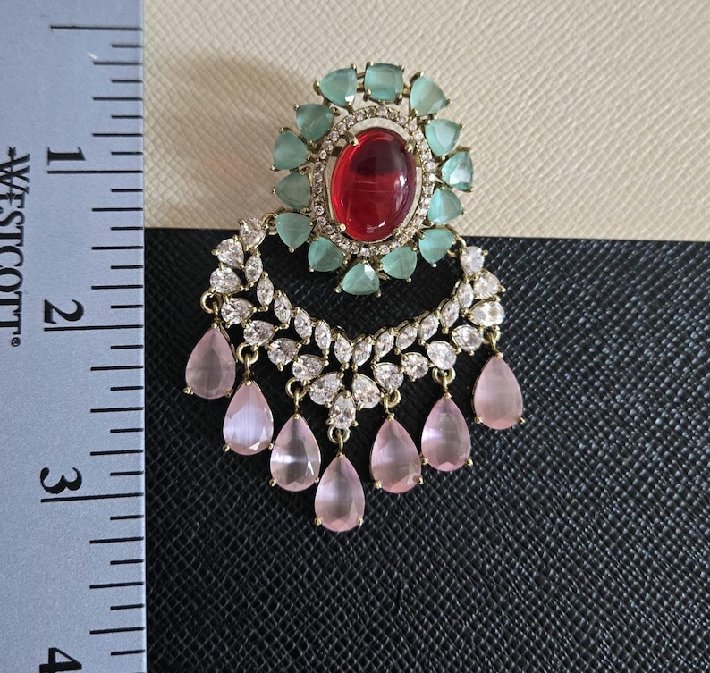 May include: An ornate earring featuring a large oval red gemstone surrounded by small, light green stones. Below, a crescent of clear, sparkling stones and dangling pink teardrop-shaped gems. The earring measures approximately 2 inches in length.