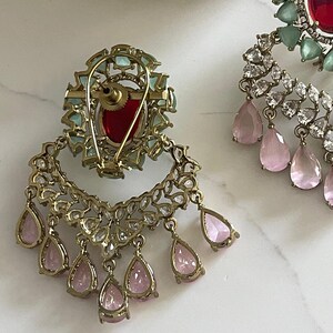 May include: Close-up of ornate gold-toned earrings. One earring features a large red gemstone surrounded by light blue stones. The other earring has a red gemstone and is adorned with clear and pink teardrop-shaped gems. The earrings are set against a white background.