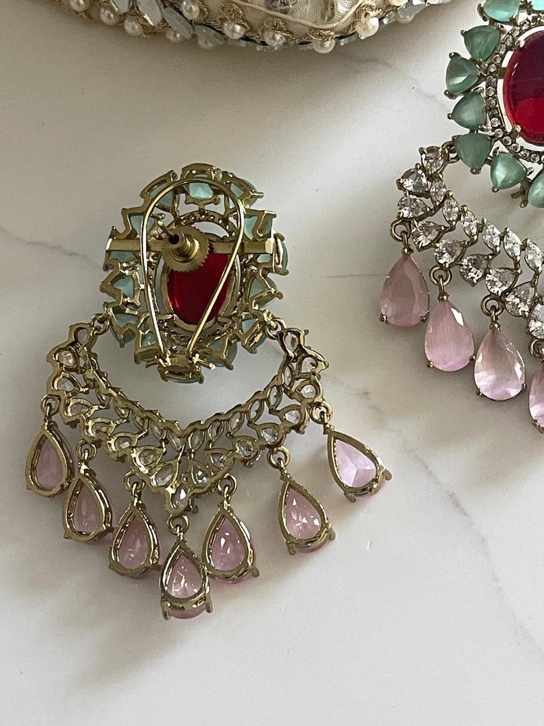 May include: Close-up of ornate gold-toned earrings. One earring features a large red gemstone surrounded by light blue stones. The other earring has a red gemstone and is adorned with clear and pink teardrop-shaped gems. The earrings are set against a white background.