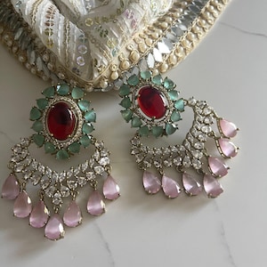 May include: A pair of ornate earrings featuring a large oval red gemstone surrounded by small green stones. Below, a crescent of clear stones and dangling pink teardrop-shaped gems. A white and gold patterned fabric is in the background.