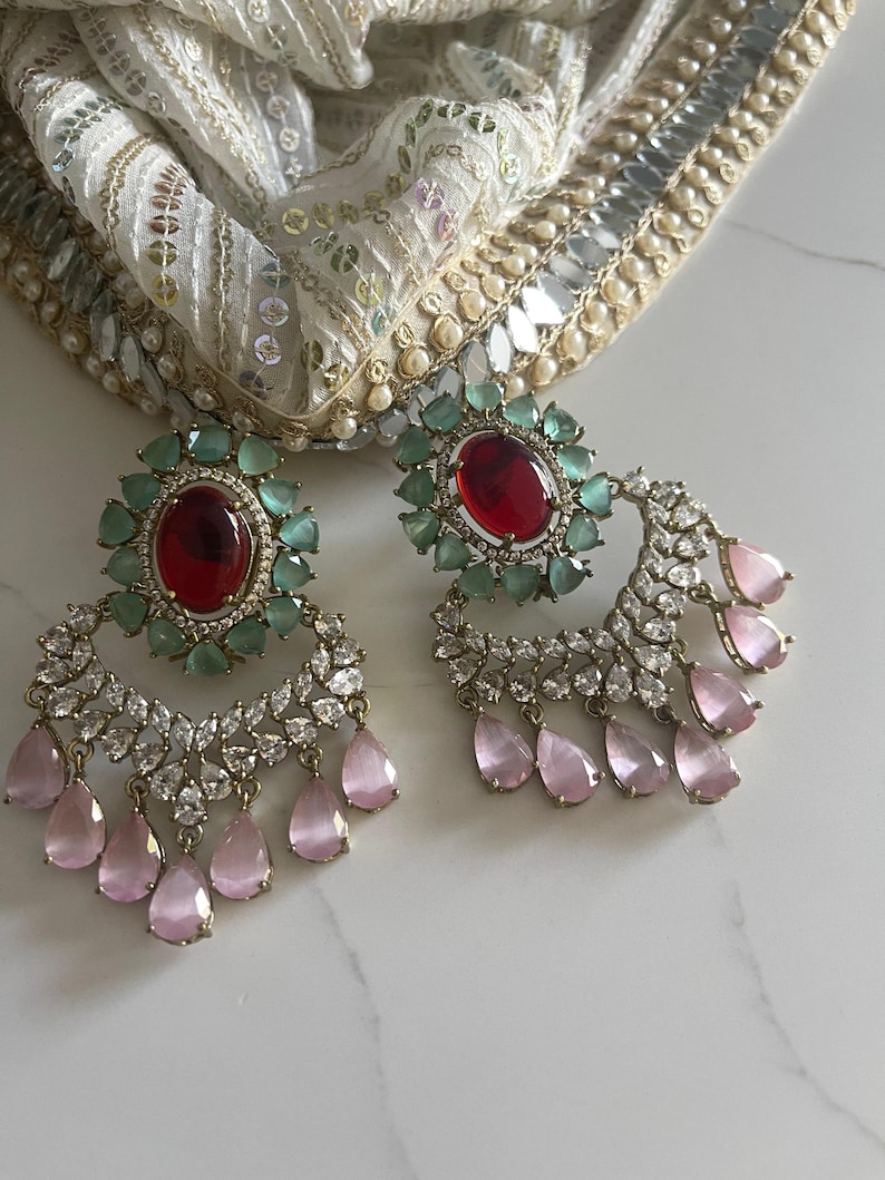 May include: A pair of ornate earrings featuring a large oval red gemstone surrounded by small green stones. Below, a crescent of clear stones and dangling pink teardrop-shaped gems. A white and gold patterned fabric is in the background.
