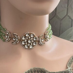 Op de afbeelding: Een gouden chokerketting met een grote ronde hanger met een bloemmotief van heldere stenen. De ketting is gelaagd met een groene kralenstreng.