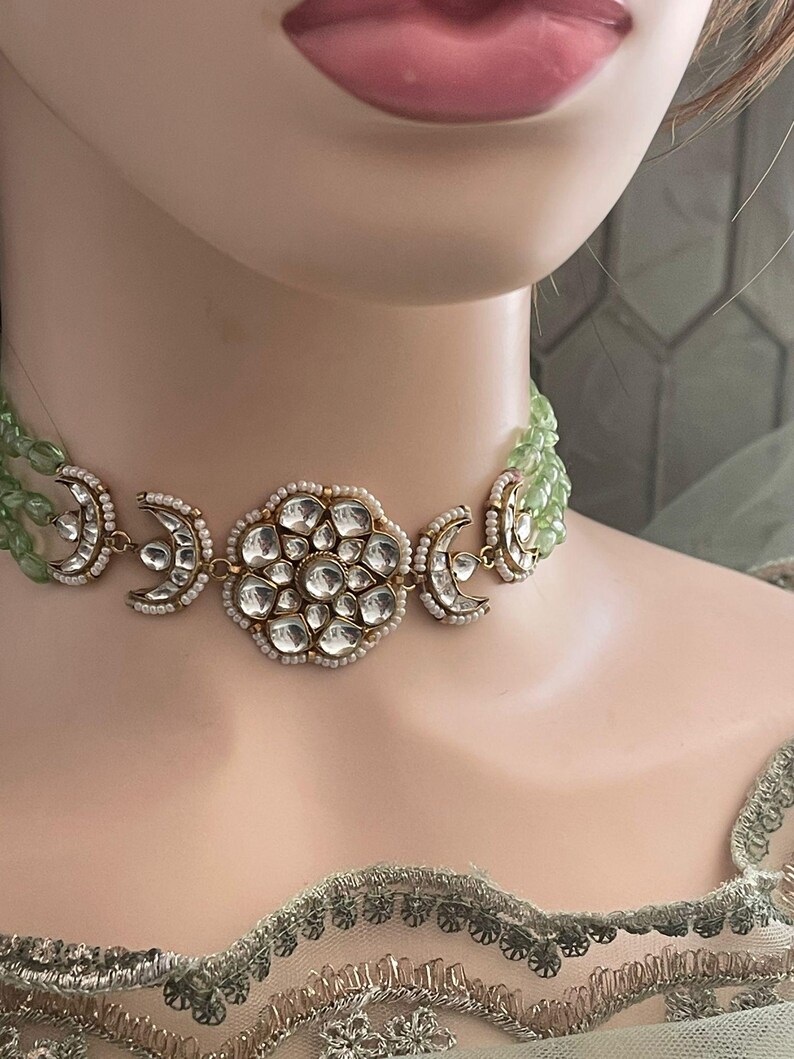 Op de afbeelding: Een gouden chokerketting met een grote ronde hanger met een bloemmotief van heldere stenen. De ketting is gelaagd met een groene kralenstreng.