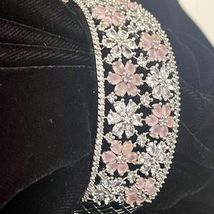 May include: A silver-toned bracelet with a floral design. The bracelet is adorned with alternating pink and clear crystal flowers. The flowers are set against a silver band, creating a sparkling and elegant accessory.