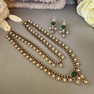 Puede incluir: Un collar de tono dorado con un colgante de piedra verde y detalles de perlas blancas. El collar es de capas y tiene un dise&ntilde;o delicado. Tambi&eacute;n se incluyen pendientes a juego con piedras verdes y perlas blancas.