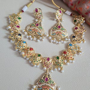 May include: A gold-coloured necklace and earring set. The jewellery features colourful gemstones, white pearls, and intricate filigree designs. The necklace and earrings have a matching design, with a central pendant and dangling elements.