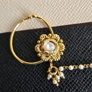 May include: A gold-toned nose ring featuring a circular hoop and a central floral-shaped ornament with a single white stone. A small cluster of dangling white beads hangs from the base of the flower, connected to a chain of small white beads.