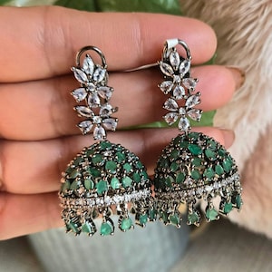May include: A pair of jhumka earrings featuring a bell-shaped design adorned with numerous teardrop-shaped green stones and clear white crystals. The top section consists of a floral-inspired crystal arrangement, with small green stone dangles.