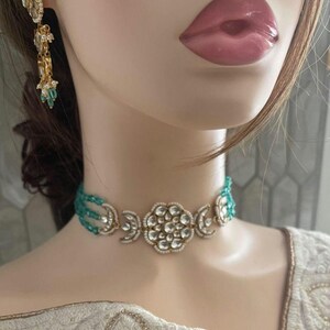 Op de afbeelding: Een gouden chokerketting met een groot bloemmotief met witte stenen en een turquoise parelrand.
