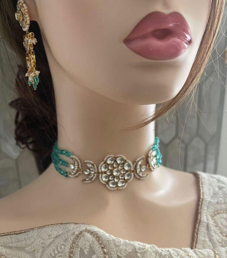 Op de afbeelding: Een gouden chokerketting met een groot bloemmotief met witte stenen en een turquoise parelrand.