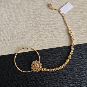 May include: A gold-toned nose ring featuring a circular hoop connected to a delicate chain. The design includes a central floral-shaped ornament embellished with small clear stones, and the chain is accented with a series of evenly spaced clear stones.
