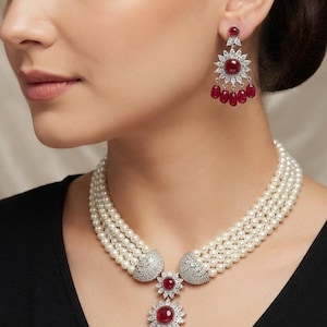 May include: A multi-strand pearl necklace featuring a central floral-shaped pendant with a red cabochon stone and dangling red teardrop beads, accented by clear crystals. Matching earrings feature a similar floral design with red stones.