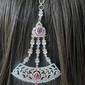 May include: A gold-toned and silver-toned headpiece with a teardrop-shaped ruby center stone and dangling accents. The headpiece features intricate floral and leaf designs, with a clip for securing it in the hair.