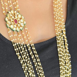 May include: A multi-strand necklace featuring small white beads and gold-colored chain. The necklace has a central pendant with a floral design, white stones, a red center stone, and green accents. The necklace is worn against a black top.