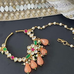 May include: A gold-toned nose ring with a floral design. The ring features clear, pink, and green gemstones, along with peach-colored teardrop-shaped beads. A delicate chain with small white pearls extends from the ring.