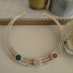 May include: A gold-toned choker necklace with three parallel bands. The necklace features three circular gemstone pendants: a green, a pink, and a rose-colored stone, each surrounded by small, clear crystals. The necklace is designed for the neck.