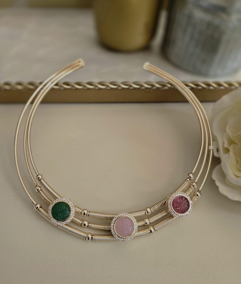 May include: A gold-toned choker necklace with three parallel bands. The necklace features three circular gemstone pendants: a green, a pink, and a rose-colored stone, each surrounded by small, clear crystals. The necklace is designed for the neck.