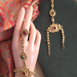 May include: Gold-toned hand chain jewelry with red and green gemstone accents. The jewelry includes a ring, a bracelet, and connecting chain. The design features square and floral elements. The background includes a patterned fabric.