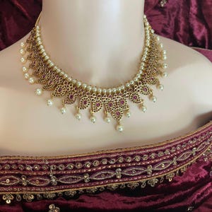 May include: A gold necklace with a pearl and ruby design. The necklace features ornate, leaf-shaped pendants with ruby-coloured stones and small, dangling pearls. The necklace is set against a burgundy background.
