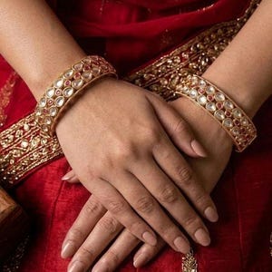 May include: Gold-toned bangles with clear and red gemstones. The bangles are worn on both wrists, set against a red fabric background with gold embroidery. The gemstones are set in a repeating pattern.