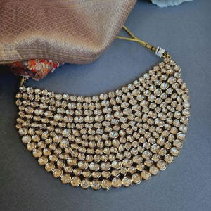 May include: A gold-coloured statement necklace featuring many clear, round gemstones. The necklace has a curved shape and is set against a dark background. A patterned fabric is visible.