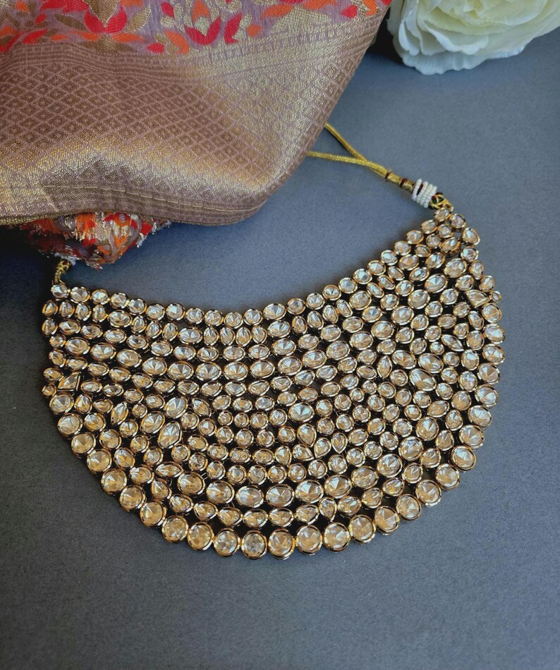 May include: A gold-coloured statement necklace featuring many clear, round gemstones. The necklace has a curved shape and is set against a dark background. A patterned fabric is visible.
