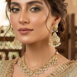 May include: A woman adorned with gold jewellery: a necklace, earrings, and a headpiece. The necklace and earrings have detailed patterns with small, pale beads. She is wearing a light green garment with gold trim.