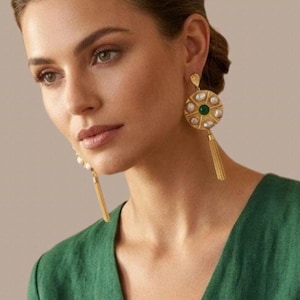 May include: Gold-toned earrings with a circular design, featuring pearls and a central green stone. The earrings have a tassel and are worn by a person wearing a green top. The earrings are a statement piece.