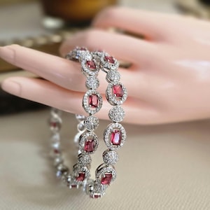 May include: Two silver bracelets with alternating ruby-red rectangular and round, clear-stone accents. The bracelets are displayed on a light-toned surface, showcasing the jewelry's sparkle and design. The bracelets are a beautiful accessory.