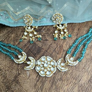 Op de afbeelding: Een turquoise kralenketting met een gouden hanger met een groot, wit, bloemmotief en twee halve manen. De ketting wordt geleverd met bijpassende oorbellen met een vergelijkbaar ontwerp.