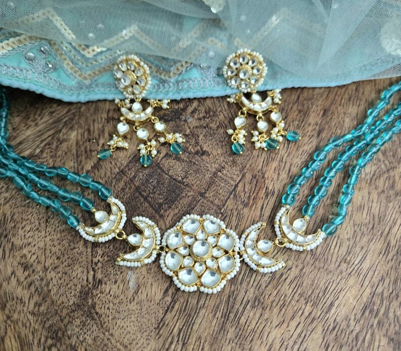 Op de afbeelding: Een turquoise kralenketting met een gouden hanger met een groot, wit, bloemmotief en twee halve manen. De ketting wordt geleverd met bijpassende oorbellen met een vergelijkbaar ontwerp.