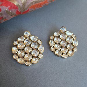 May include: A pair of gold-coloured stud earrings. Each earring has a cluster of clear, faceted stones in various shapes, set in a circular arrangement. The earrings are displayed on a grey surface, with patterned fabric in the background.